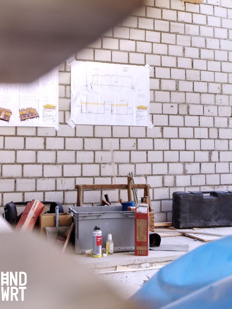 Baustelle mit weißer Fliesenwand, Bauplänen und Werkzeugkoffern auf hellem Estrichboden