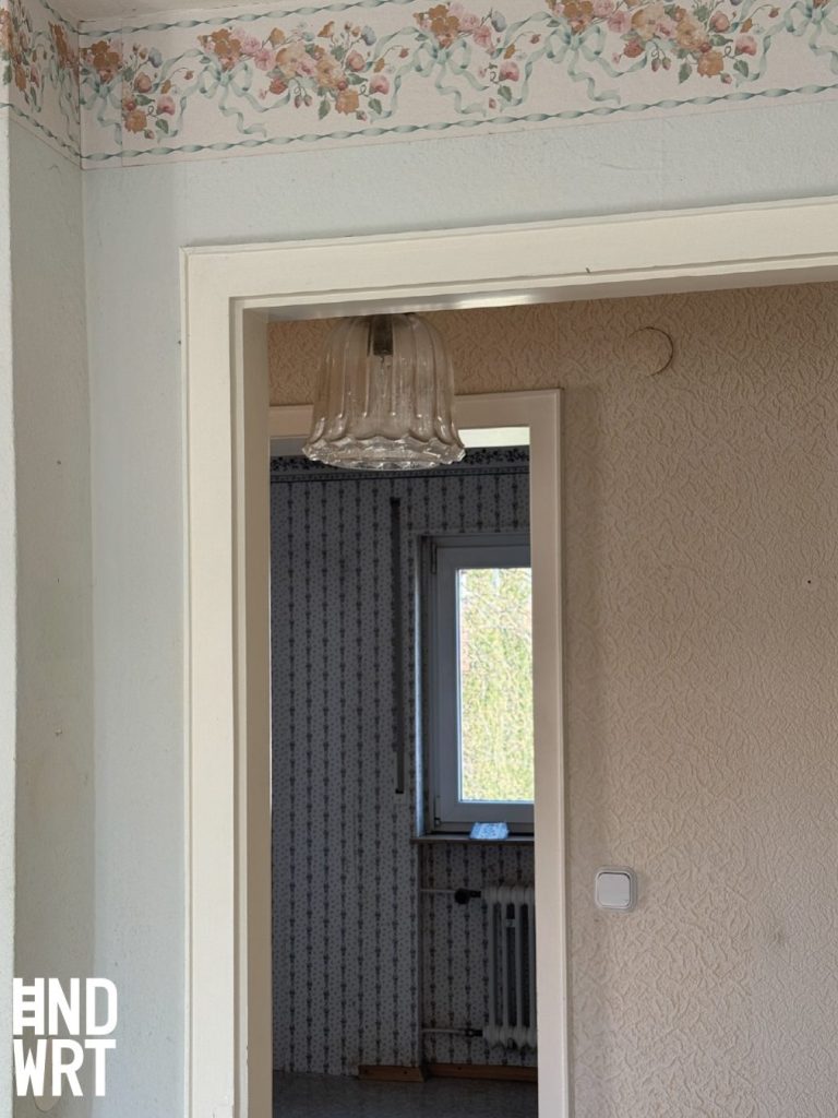 Altbau-Innenraum mit Blumentapete an der Decke, weißer Türzarge und Blick in Raum mit Raufasertapete und Glasleuchte