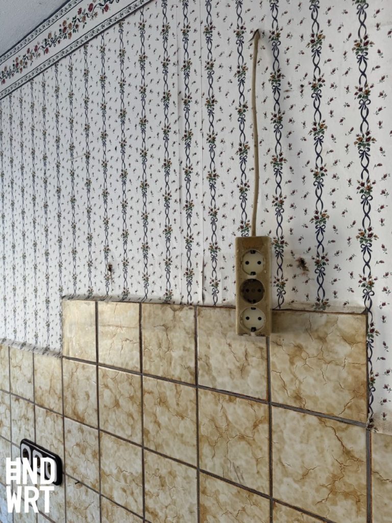 Küchenwand mit beigefarbenen Wandfliesen und Blumentapete, Steckdosen teilweise montiert, Sanierung im Gange