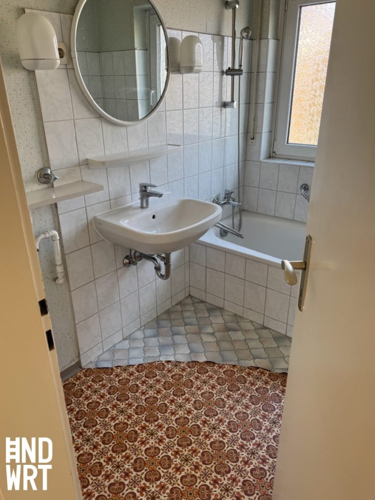 Badezimmer mit weißen Wandfliesen, Waschbecken, Badewanne, Duschkabine und gemustertem Fliesenboden im Eingangsbereich