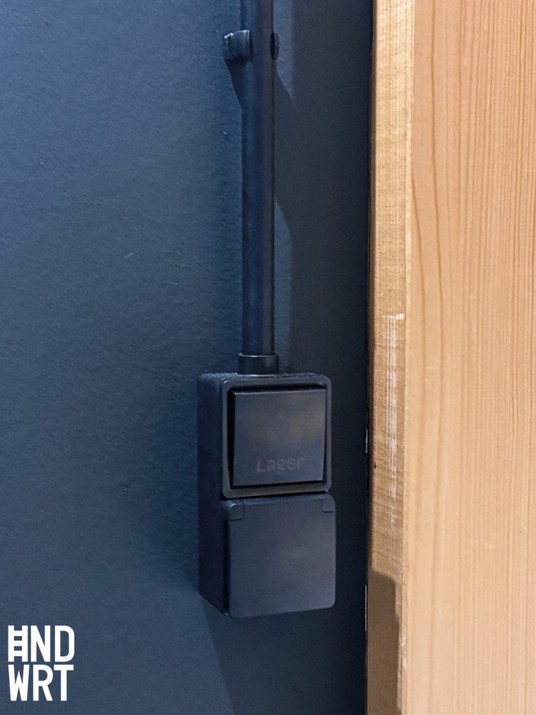 Schwarze Aufputz-Steckdose mit Lichtschalter an dunkelblauer Wand neben hellem Holztürrahmen