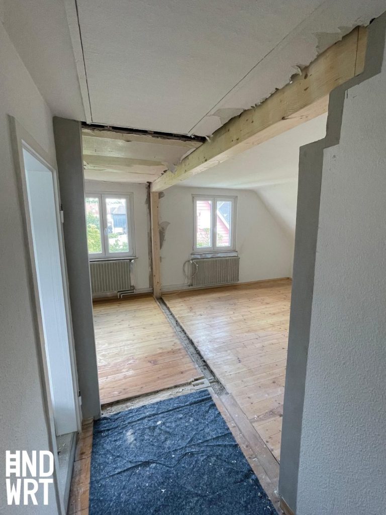 Raum mit teilweise abgebrochener Decke, freiliegenden Holzbalken, Holzdielenboden und weißen Wänden während Abbrucharbeiten