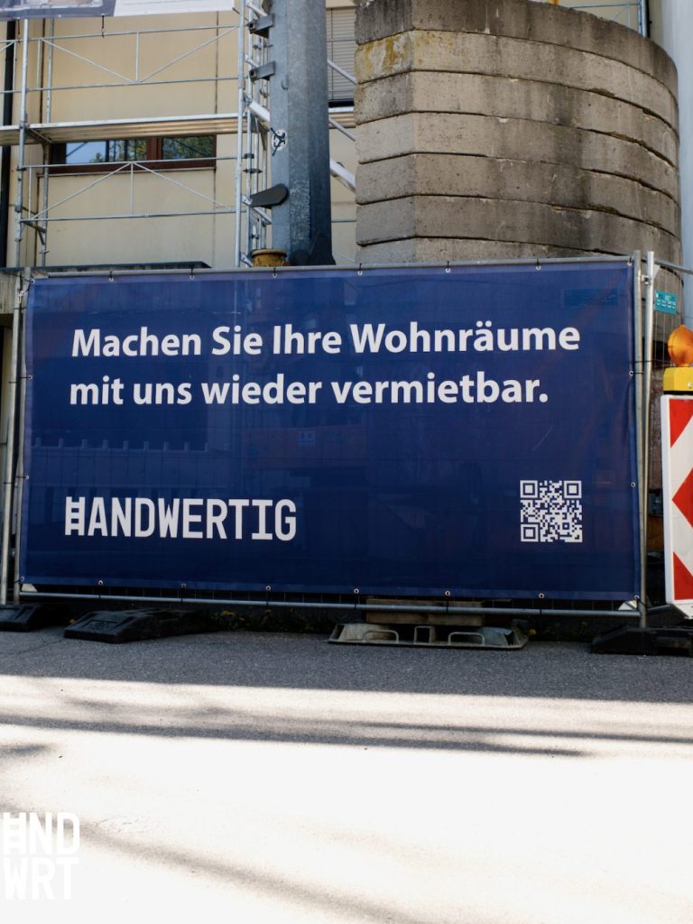 Baustelle mit Gerüst, Betonsiloturm und blauem Bauzaun mit Werbebeschriftung und QR-Code