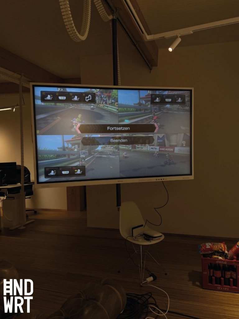 Großer Bildschirm an Deckenhalterung in Raum mit Holzboden zeigt Videospiel mit Rennstrecke und Menüoptionen